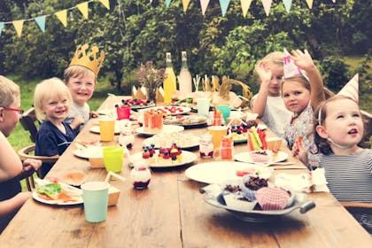 Outdoor picnic birthday party with children sat at a wooden table, wearing party hats 