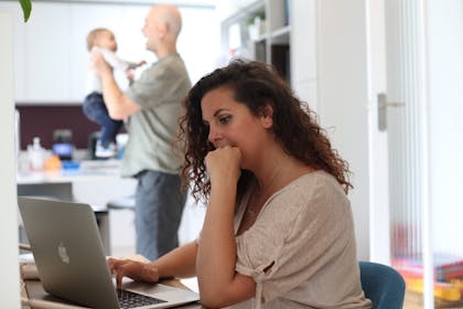 Woman working at laptop at home while man holds baby in background
