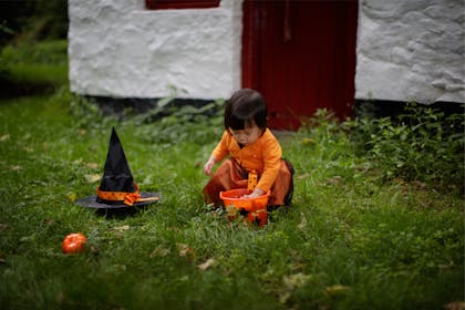 A toddler in an orange Halloween dress plays in the garden with a witch's hat and an orange bucket