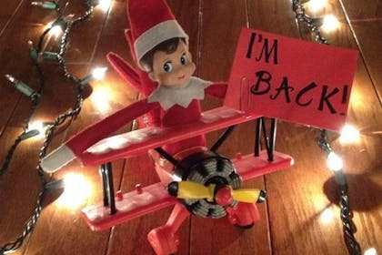 Elf on the Shelf arriving via runway fairy lights holding a sign saying 'I'm back!'