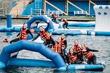 A smiling group of women wearing life vests and wetsuits wave while resting on an inflatable obstacle at Let's Go Hydro 