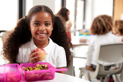 Child in school uniform with pink lunch box smiling and holding apple 