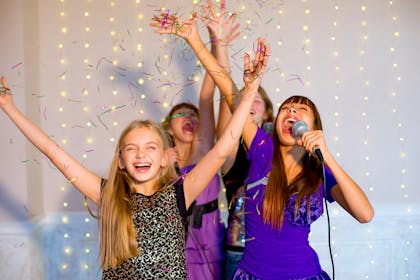 Four girls sing karaoke and throw confetti with fairy lights in the background