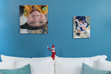 Elf on the Shelf sitting on sofa, pictures on the wall have been turned upside down