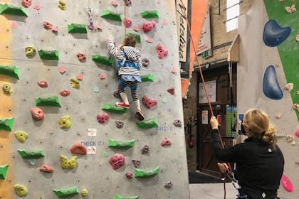 A young girl in a stripy hoody and leggings holds onto a climbing wall while a teacher watches and holds her rope.