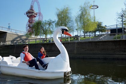 A laughing couple wearing life vests pedal a swan pedalo around the Olympic Park in London
