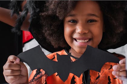 Little girl in orange Halloween costume holds up black paper bat