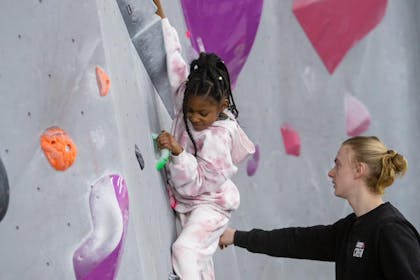 A girl in a pink tracksuit climbs at a Hangar Climbing centre with the help of a teacher
