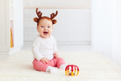 Baby wearing Christmas Rudolf antlers sitting on the floor next to a toy