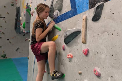A smiling girl in shorts and t-shirt clings to a climbing wall at The Climbing Unit in Derby
