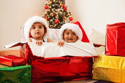 Two babies wearing Santa hats peeking out of a big red Christmas gift box