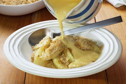 Custard poured from jug onto apple crumble in bowl