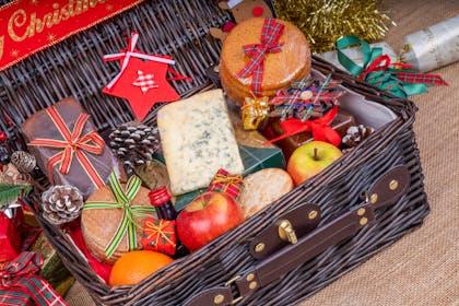 A Christmas hamper gift filled with cheeses, apples, oranges, bread, a bottle of wine and decorated with stars and pine cones
