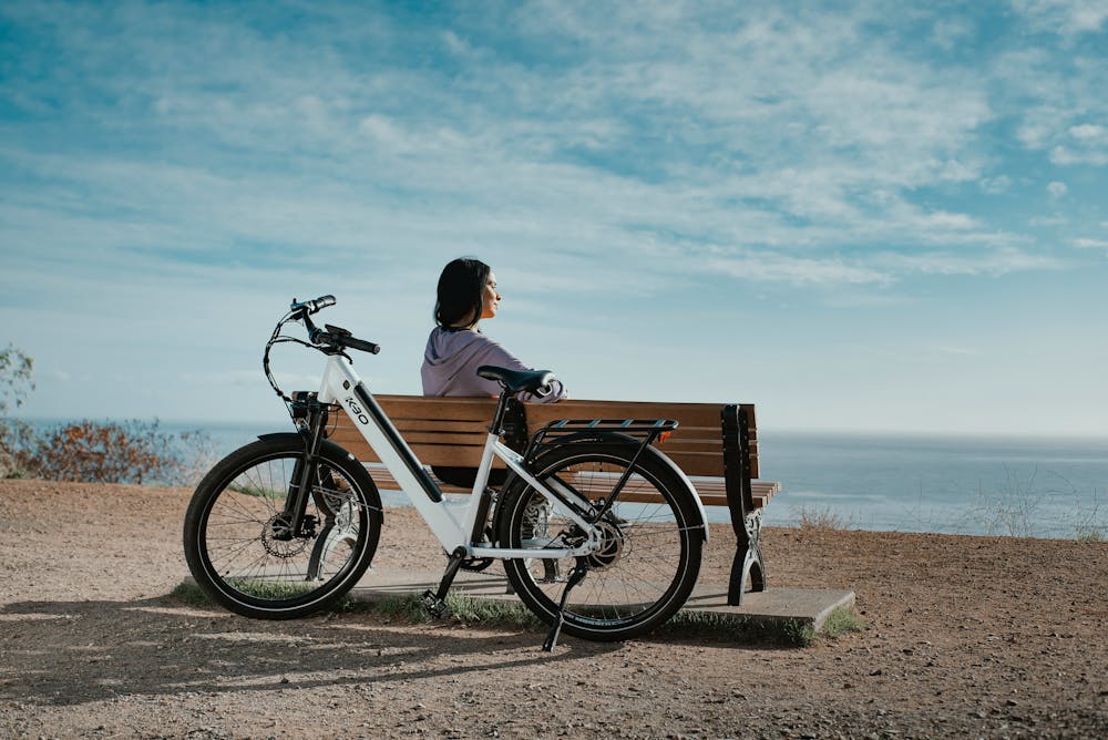 Une femme assise sur un banc en face de la mer, avec son vélo électrique reconditionné garé derrière elle