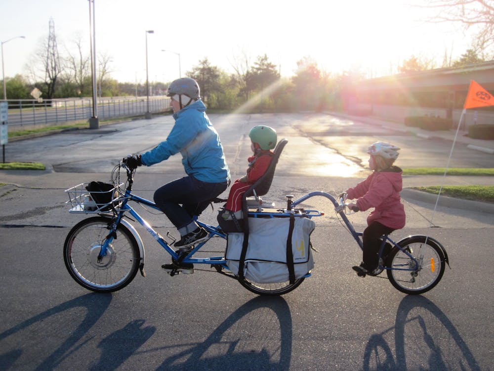 Un parent à vélo avec un bébé en porte-bébé, et un enfant en remorque vélo