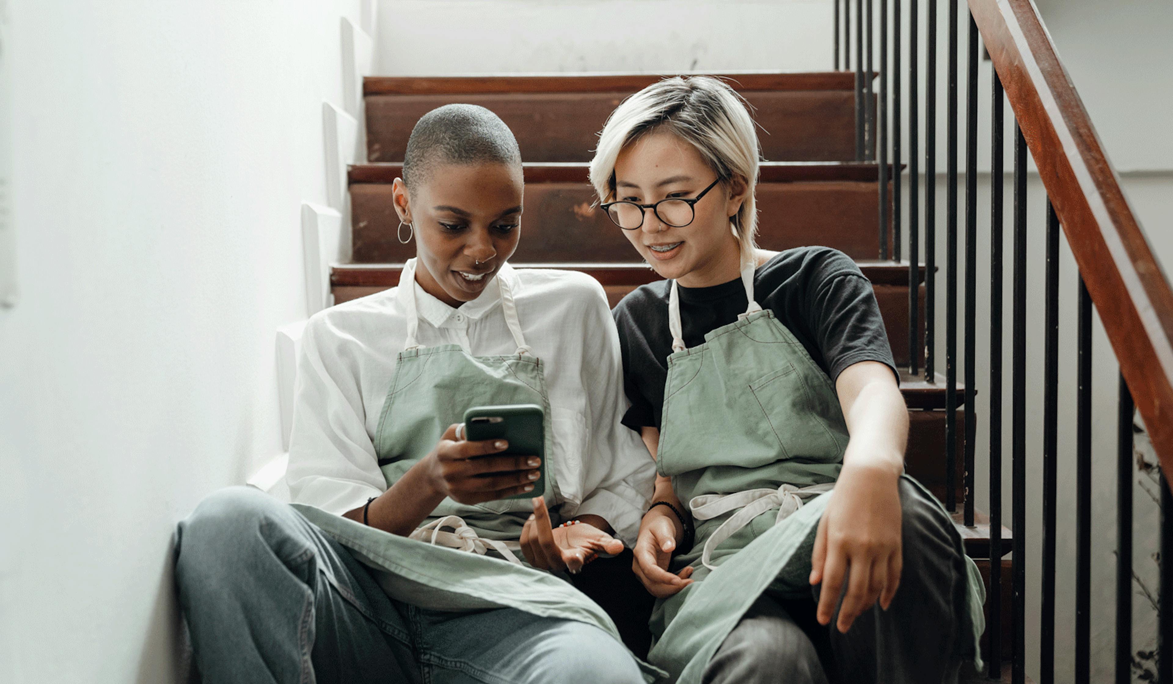 Image of two restaurant worker sitting on the stairs looking at an iPhone