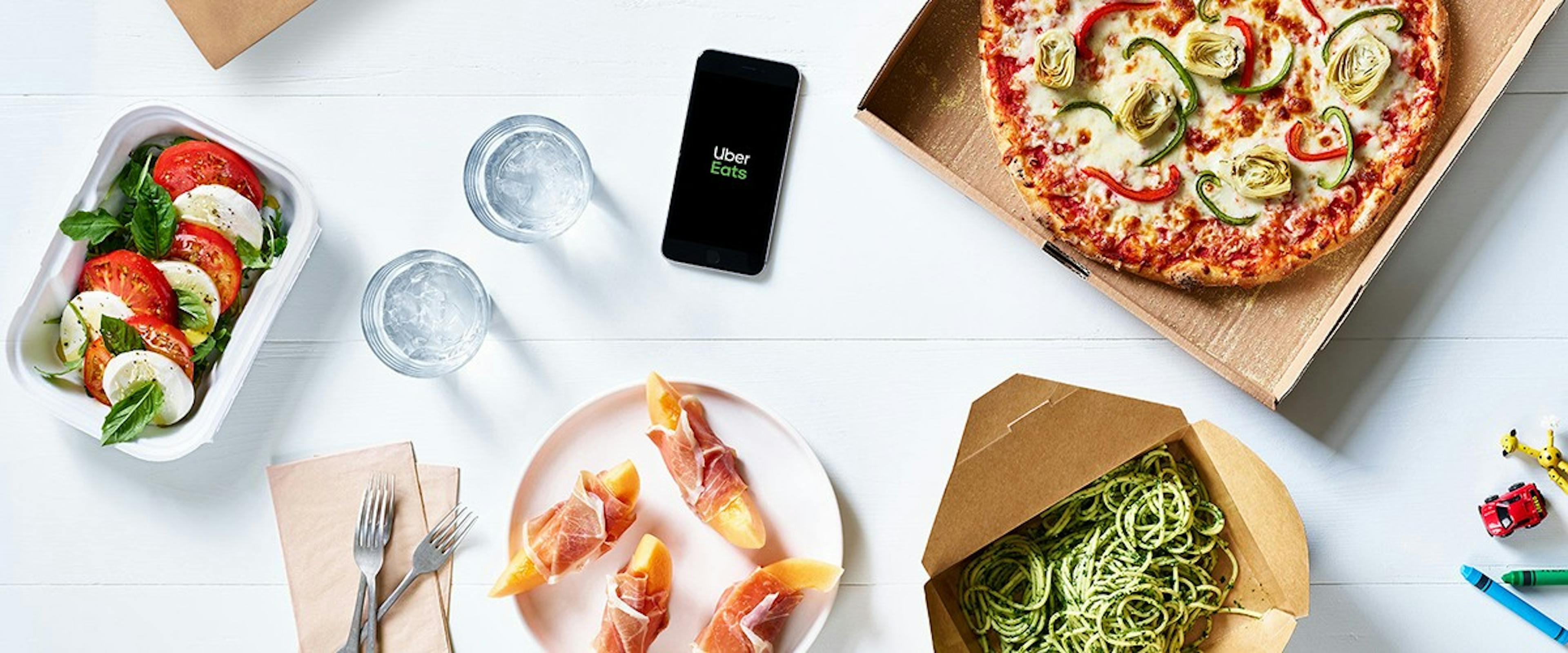 Image of table filled with food including pizza, melons & prosciutto, pasta and caprese salad with an iPhone open to Uber Eats in the middle