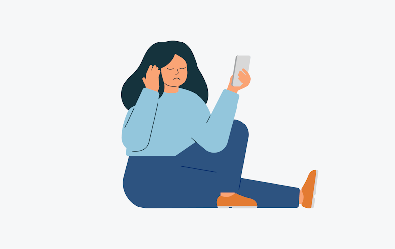 Manneskja situr flötum beinum á gólfinu með annan fótinn krossaðan yfir. Hún er leið á svip og með lokuð augun. Hún er með dökkt sítt hár, er í blárri peysu, dökkum buxum og brúnum skóm. Hún heldur hægri hendinni upp að eyranu en heldur farsímannum upp fyrir framan sig í vinstri hendinni.