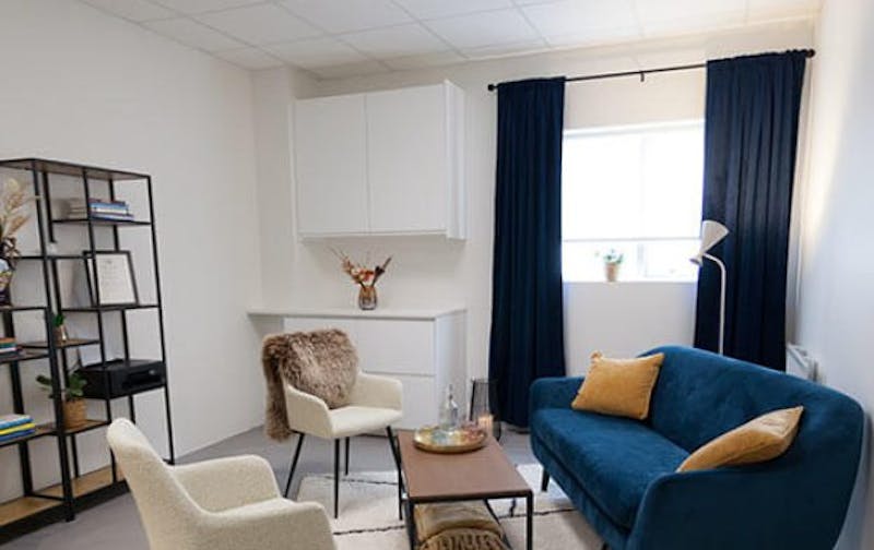 Myndin sýnir bjarta setustofu þar sem er blár sófi hægra megin upp við vegg.  Sófinn er með tveimur gulum púðum fyrir framan lágt viðar sófaborð. Hinu megin við borðið eru tveir hvítir stólar, yfir annan þeirra hefur verið lagt samanbrotið teppi.  Fyrir aftan stólana má sjá bókahillu með ýmsum munum í. Á veggnum gagnstætt hurðinni er hægra megin gluggi með bláum gluggatjöldum. Vinstra megin eru hvítir upphengdir eldhússkápar.