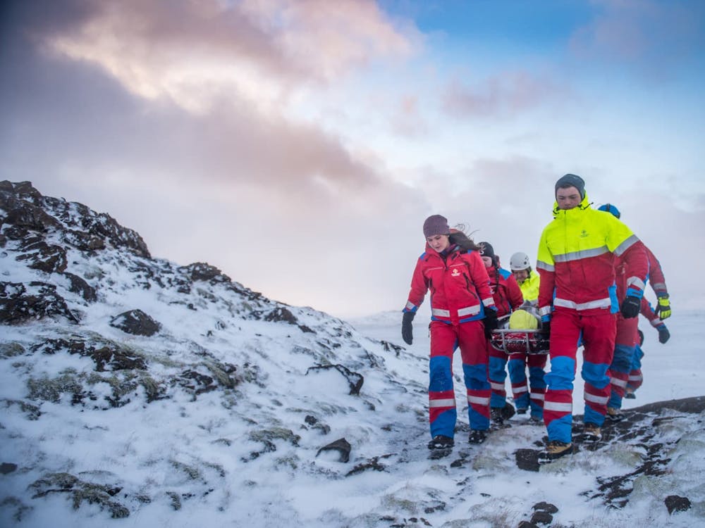 Björgunarsveitarfólk kemur gangandi yfir hæð með börur á milli sín. Það er snjór yfir og landslagið er grýtt.