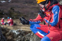 Björgunarsveitarmaður í forgrunni að tala í talstöð. Hann horfir frá okkur yfir á hóp björgunarsveitarmanna í fjarska sem krjúpa í hrauninu í kringum börur.