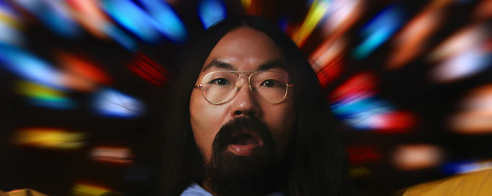 Homme barbu aux cheveux noirs longs, portant des lunettes avec des verres optiques Nikon, entouré d’un fond flou et coloré illustrant la correction visuelle.