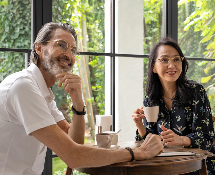 Un couple souriant, tous deux portant une paire de lunettes avec des verres optiques Nikon, buvant un café en tête-à-tête.