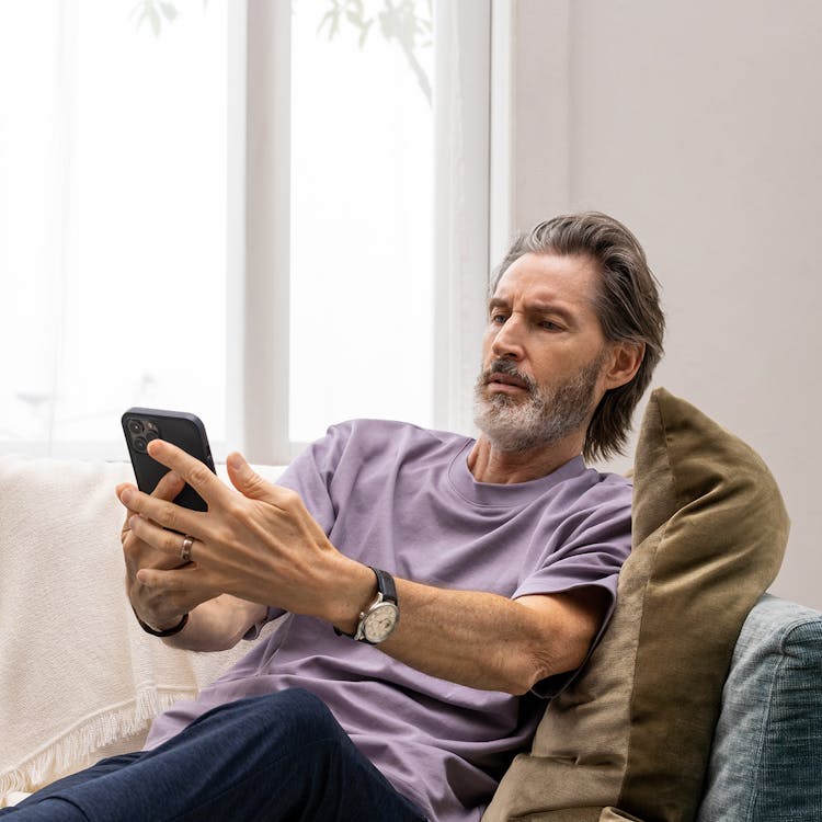 Homme ayant du mal à regarder son téléphone en fronçant ses sourcils à mi-distance assis sur son canapé.