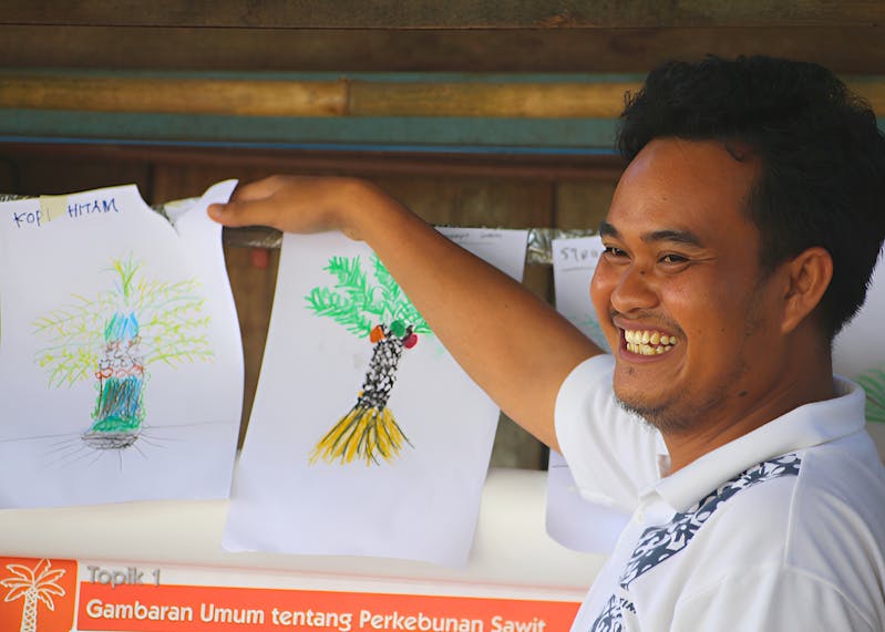 Training in Kalimantan - smiling man gestures toward colorful drawings of plants hanging on the wall.