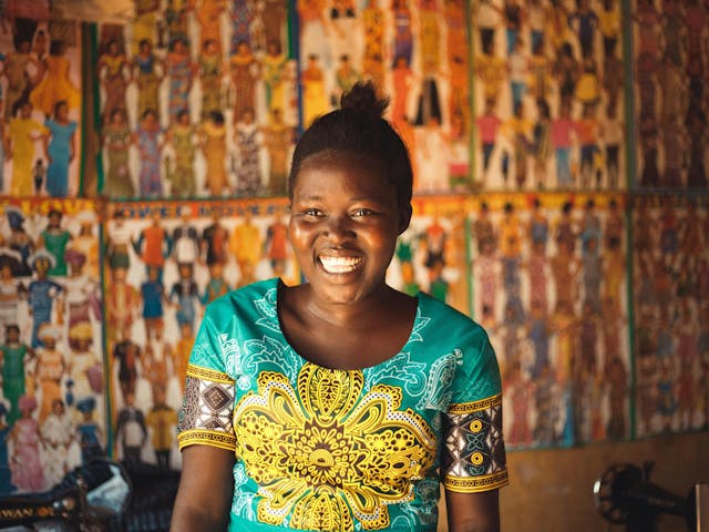 Close-up of a young Black woman with her hair pulled back, wearing a bright teal and gold patterned shirt, smiling widely. Behind her is a wall covered with many small, colorful illustrations of people in traditional clothing or designs, and a glimpse of a sewing machine on the left.
