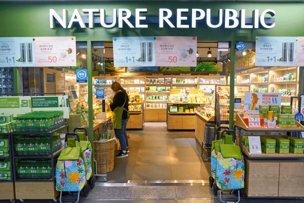 The outside of popular Korean skincare and Korean hair care brand Nature Republic, with many products outside and inside of the store and a worker inside in a green apron.