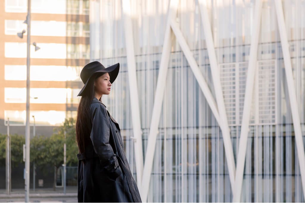 A woman stands in a stylish black pleather trench coat with a black brimmed hat on too in front of a white wall