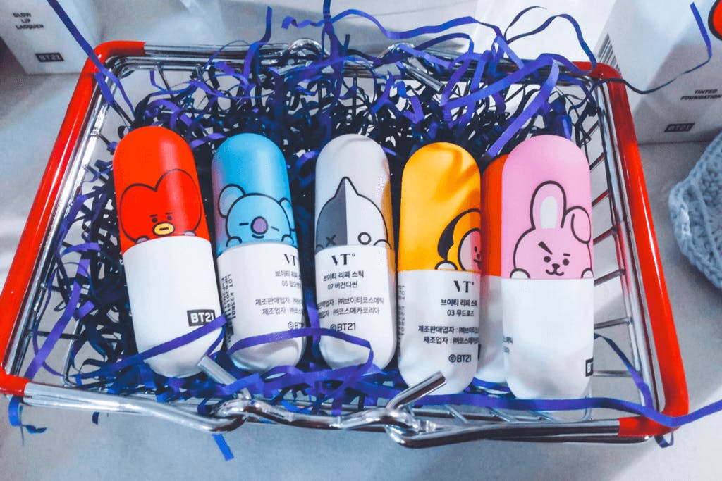 A red cart full of blue streamers and six VT BT21 makeup products with the label written in Korean with 5 different characters on the front of each.