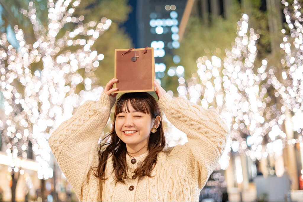 A girl holding a small shopping bag above her head with Christmas lights behind her.