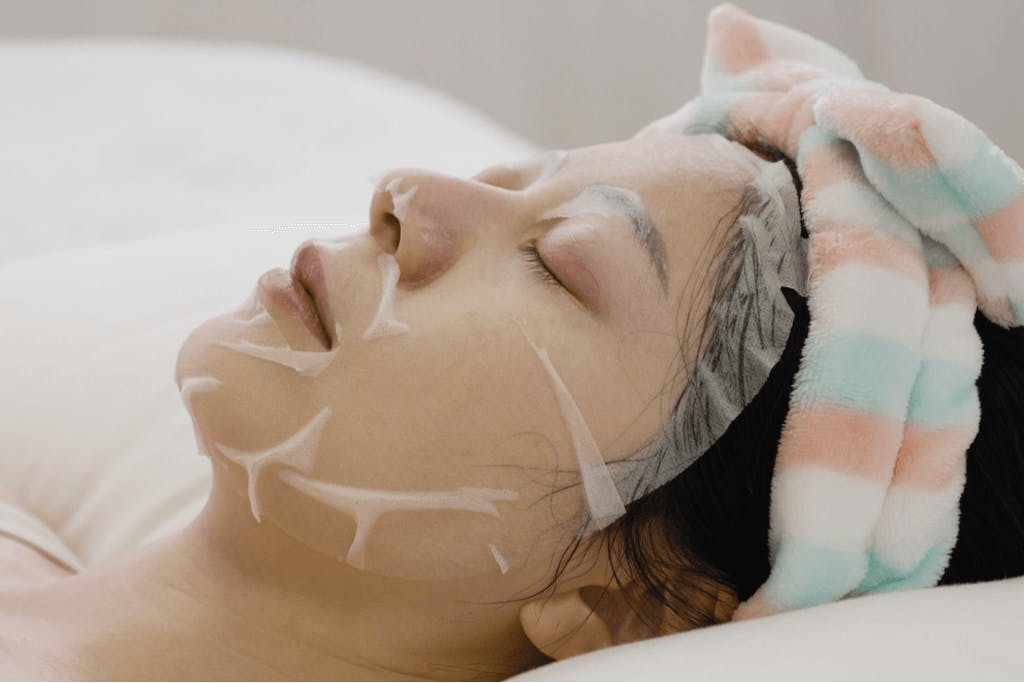 A woman rests in her bed with one of the best Korean sheet masks for her skin on her face.