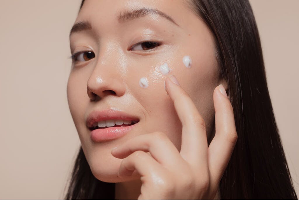 A Korean woman wipes moisturizer onto her cheek while making a coy face