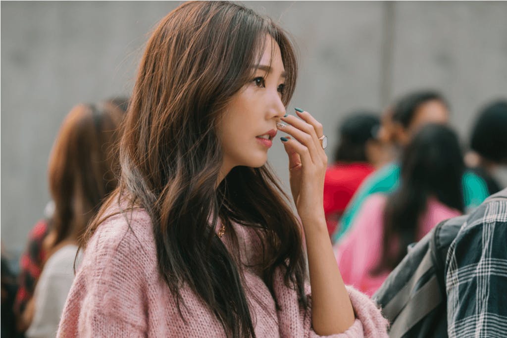 A woman brushes hair out of her face with rosy cheeks, a popular part of current Korean makeup trends, in front of a group of people.