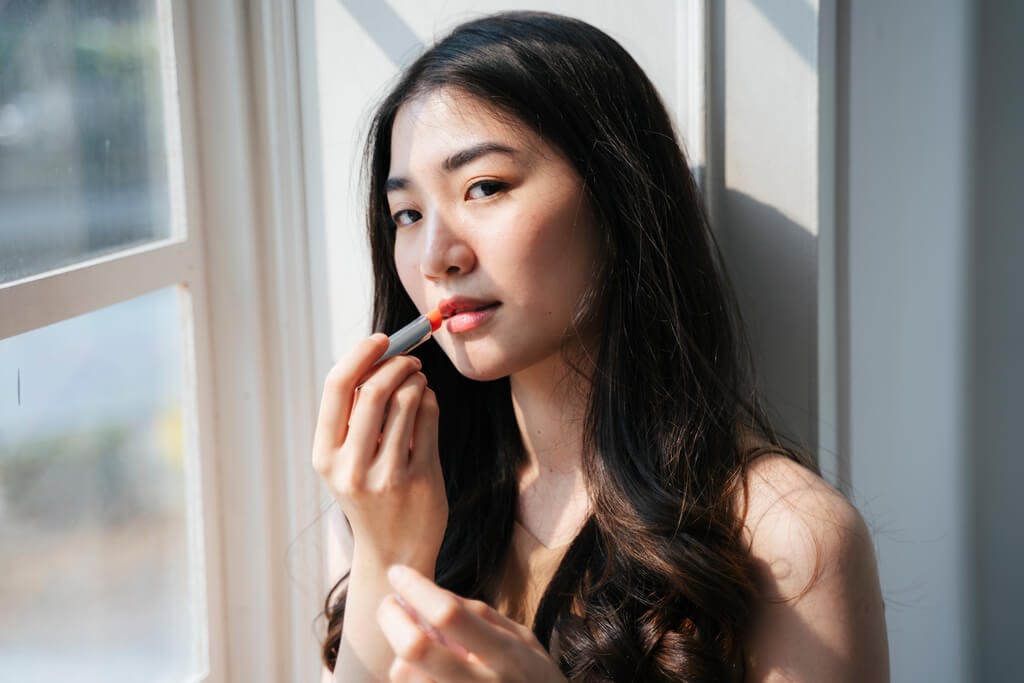 A woman stands next to a window while looking at the camera and putting on a stick-type lip tint.