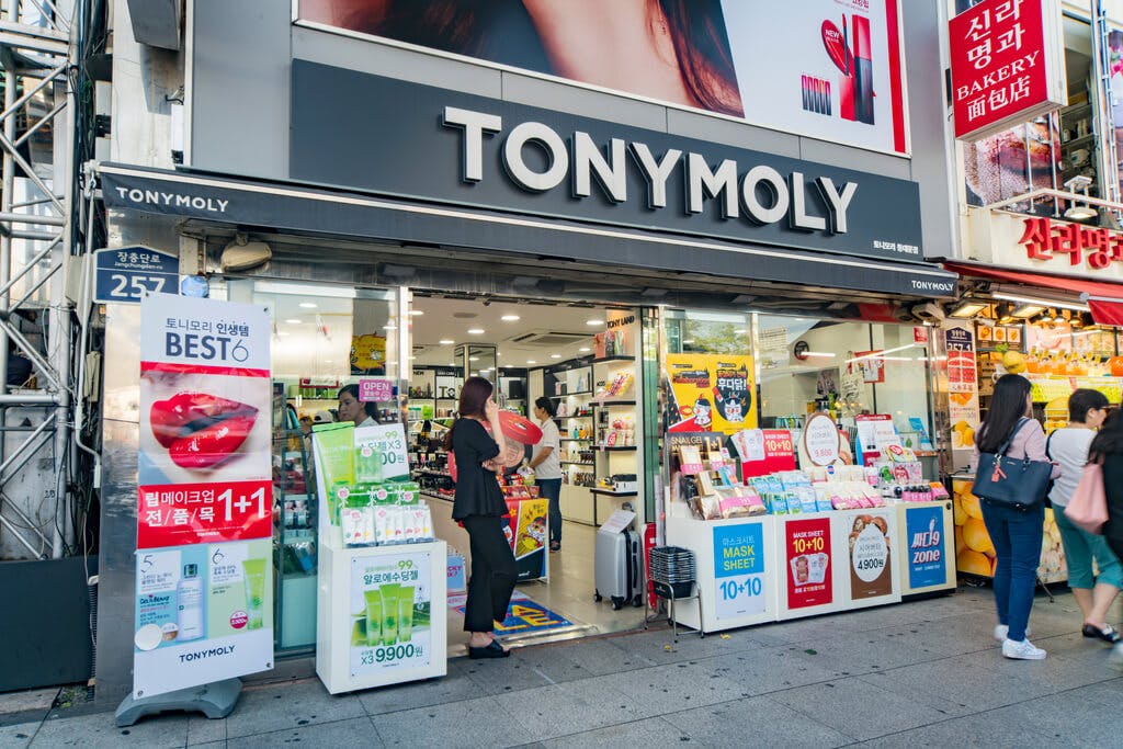 The exterior of a Tony Moly store with the doors open and people standing inside and people walking outside with several displays