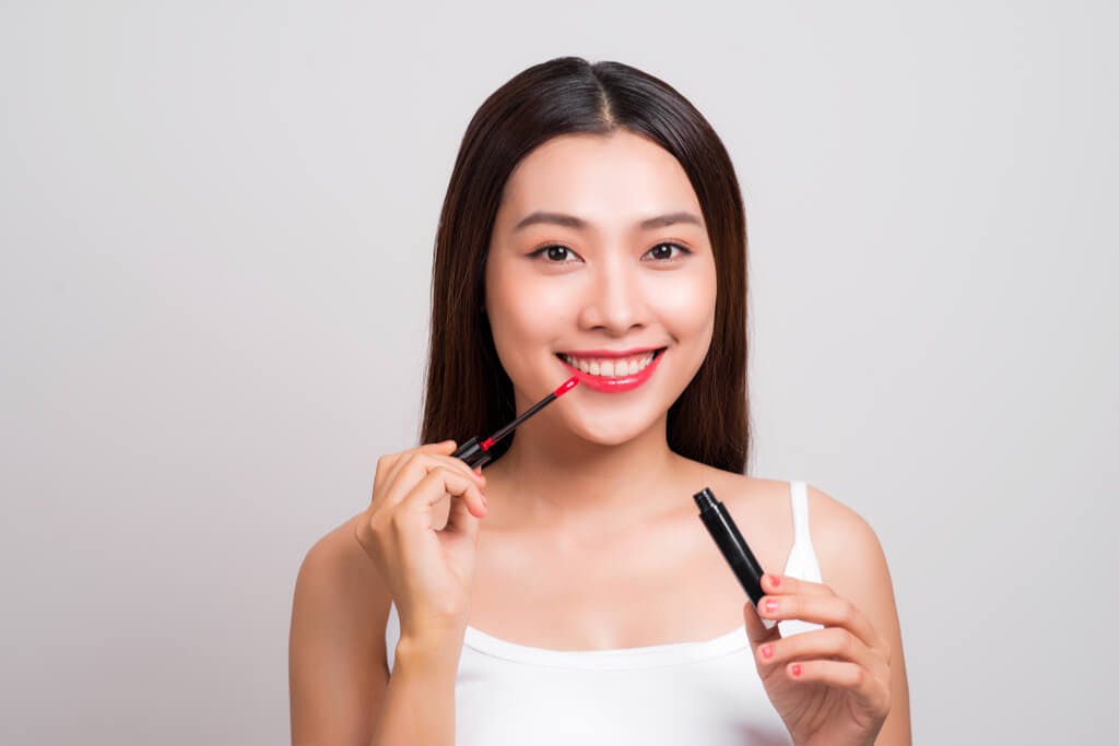 A woman smiles as she applies a bold red lip tint to her lips in front of a gray background.