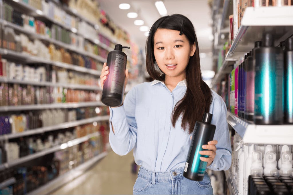 A woman holding bottles of Korean hair care products in a store with many more products on shelves behind her in a store.