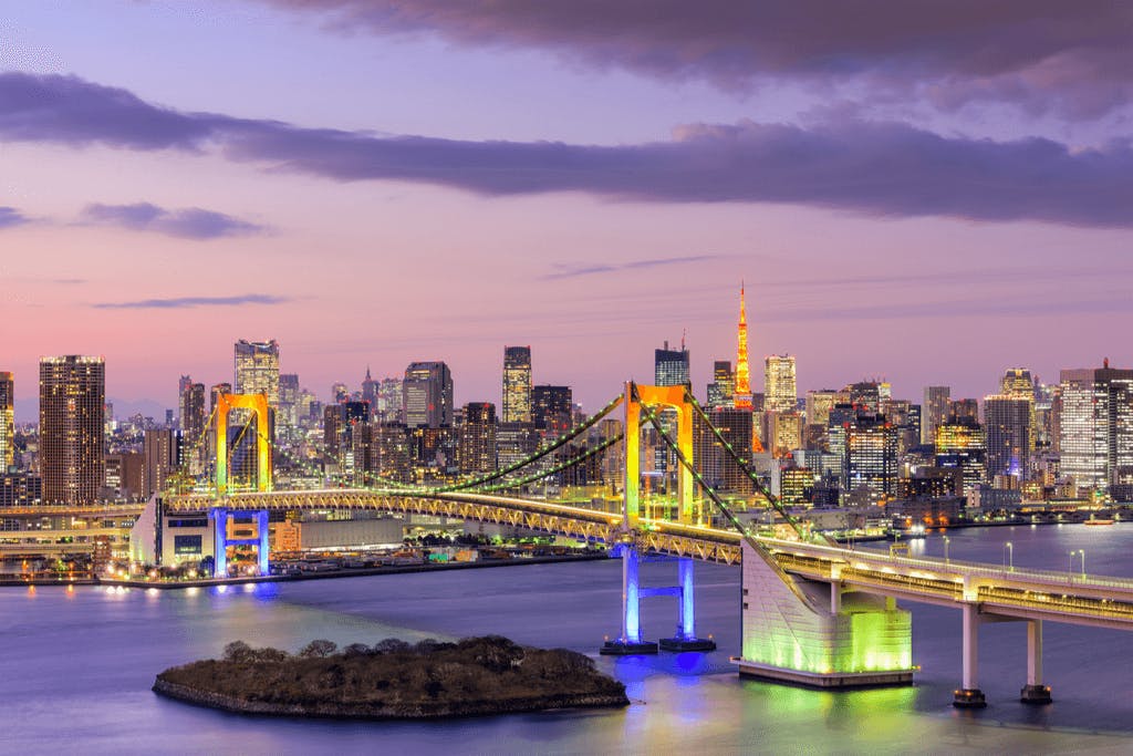 Tokyo's Rainbow Bridge with two of the arches lighted with rainbow lights and a view of the city in the background with the river flowing underneath.