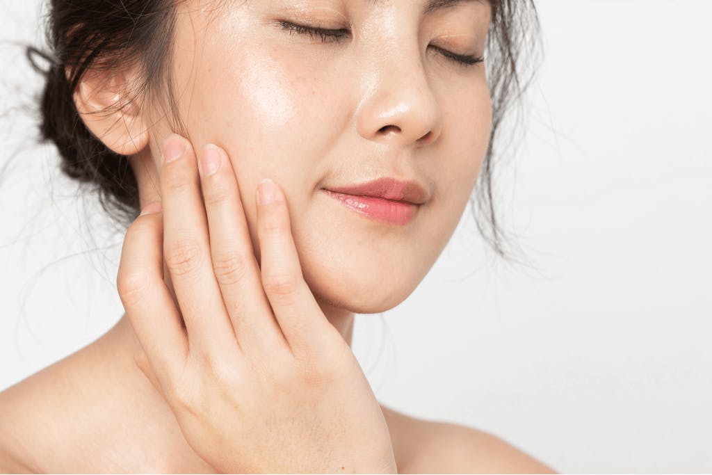 A woman with shiny glass skin, one of the current Korean makeup trends, in front of  a white background as she touches her face.