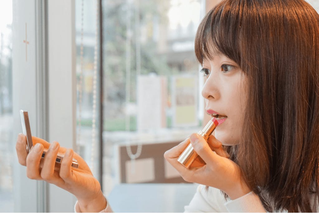 A woman in a store applying lipstick with one hand and holding a compact mirror in the other.