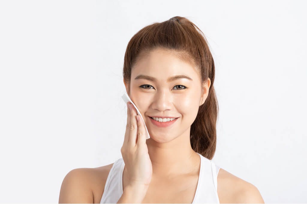 A young woman wipes Korean toner onto her face in front of a white background