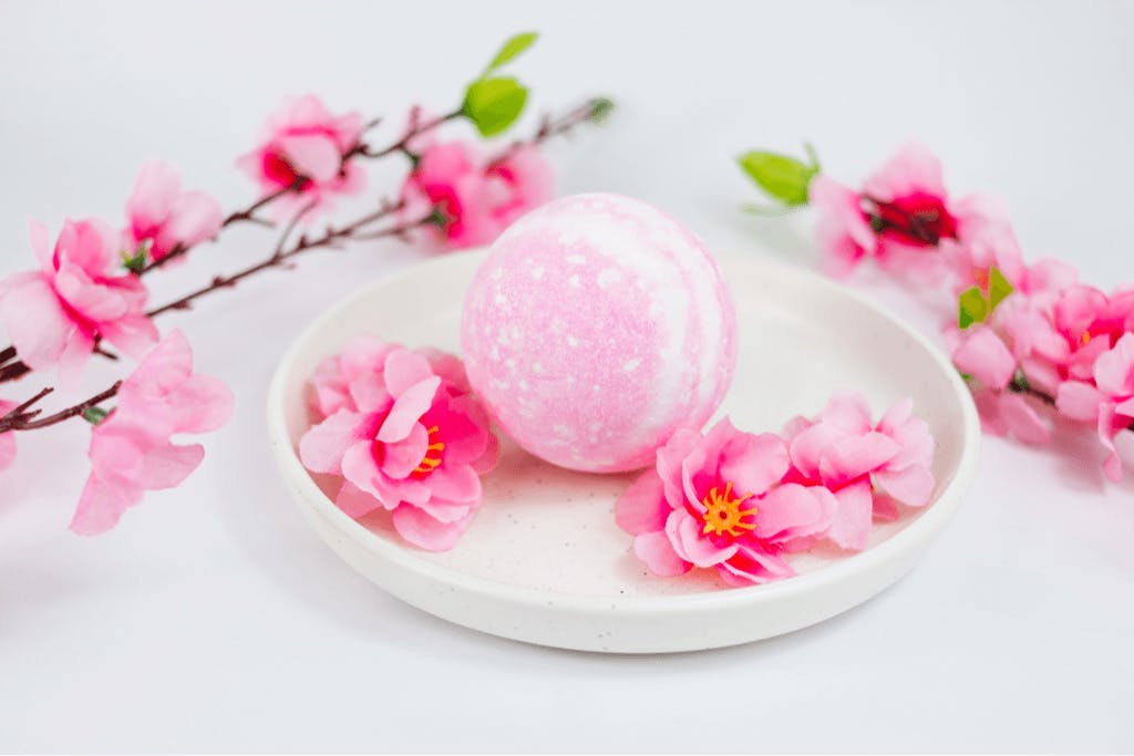 A sakura-themed and scented Japanese bath bomb sits on a white plate with cherry blossoms around it both on the plate and off of it.