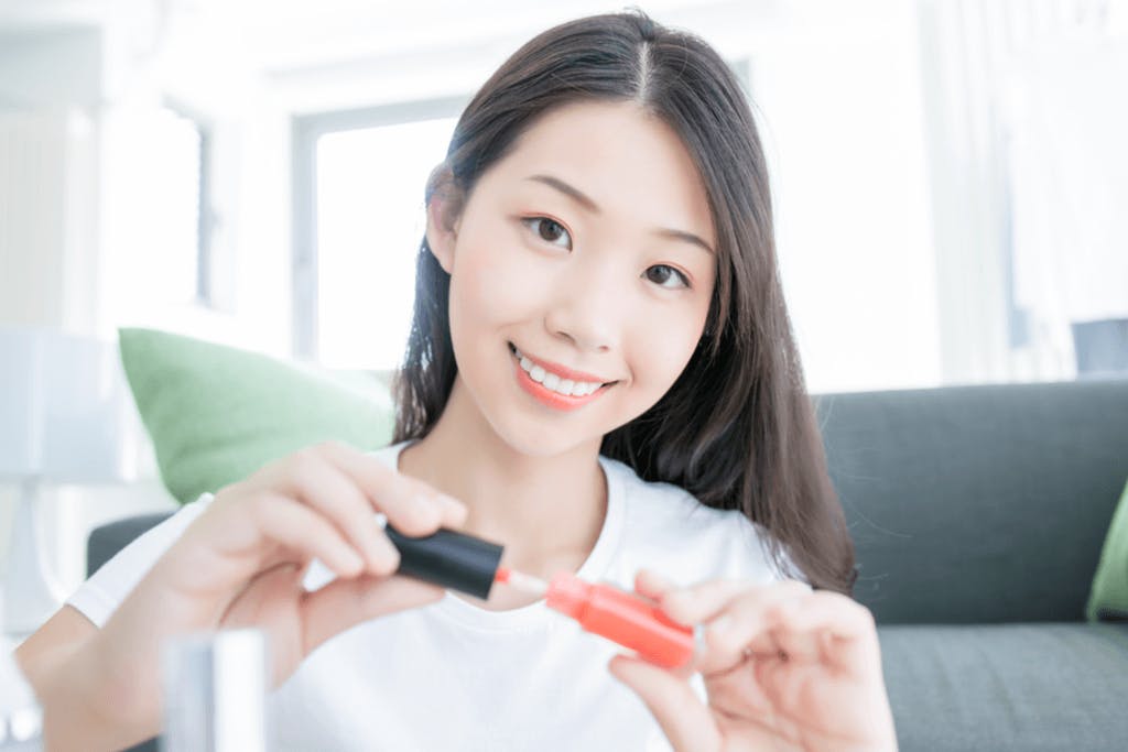 A woman smiles at the camera as she removes the applicator from a bottle of Korean lip gloss in her living room with a comfy sofa behind her