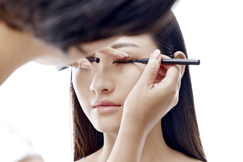 A woman sits in a chair as she gets her eyeliner drawn on by a makeup artist in front of a white background.