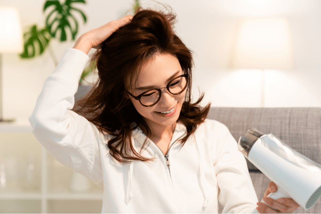 A woman in glasses dries her hair with a hair dryer while running her hands through part of it.