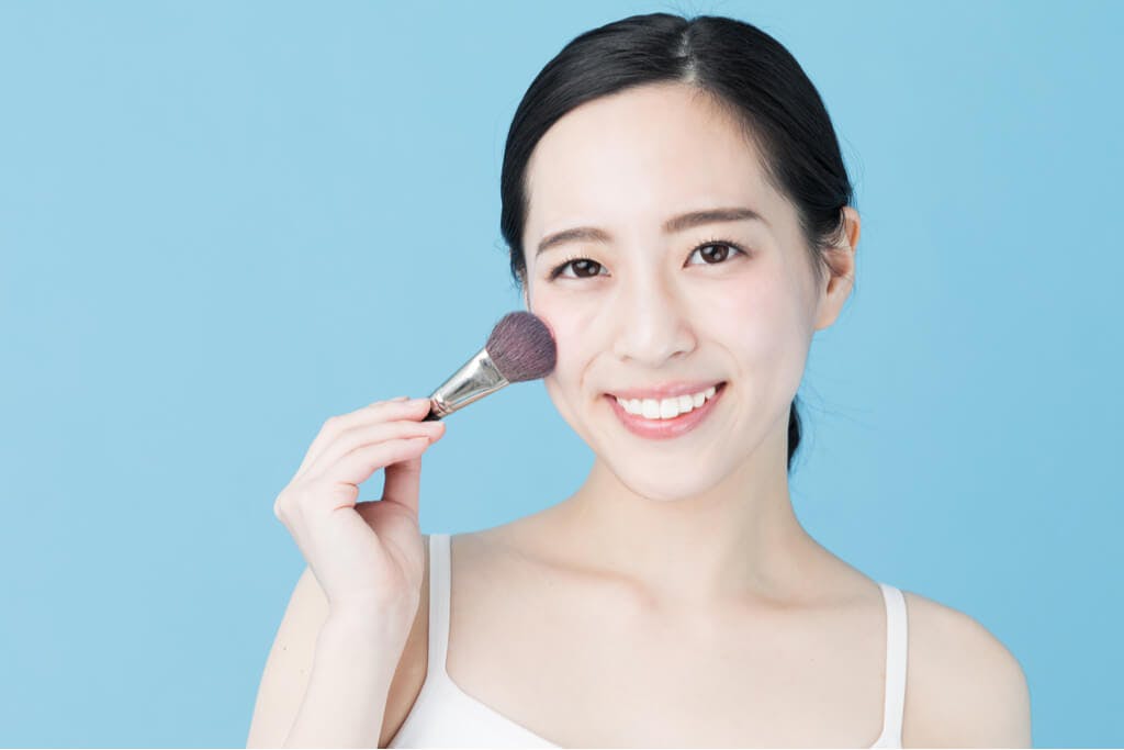 An Asian woman in front of a blue background holding a foundation brush to her skin.